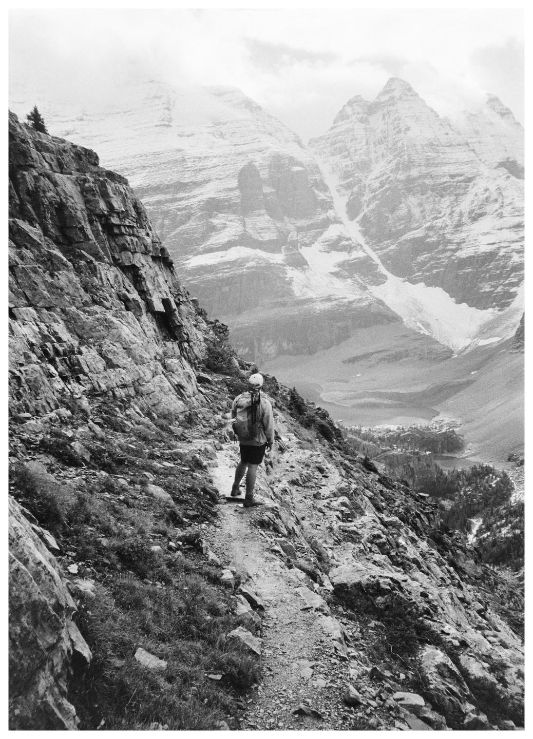 A person with a backpack walks along a narrow mountain trail overlooking a valley with a lake and distant mountains.