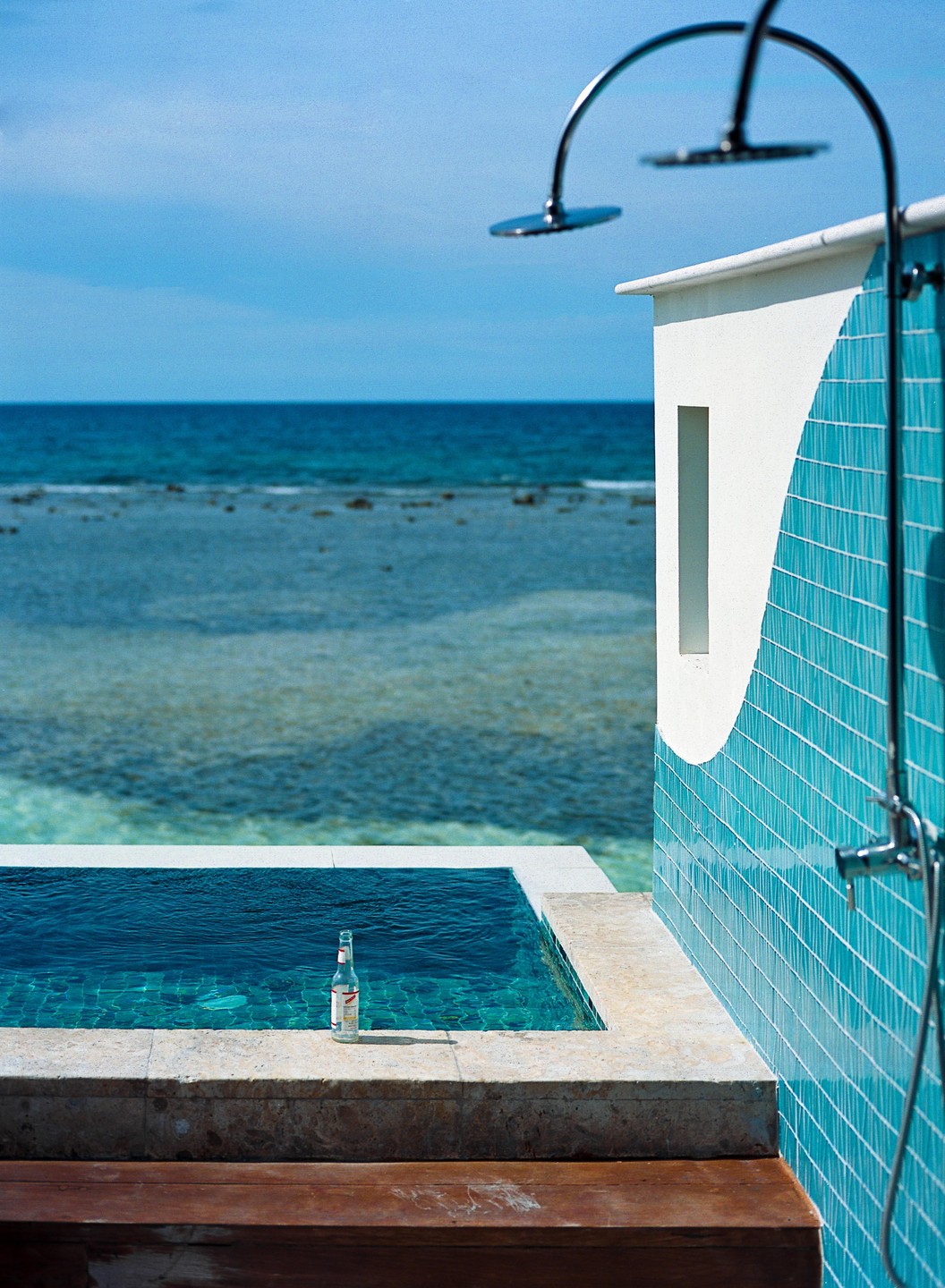 A bottle stands on the edge of a small pool next to a tiled wall with showers, overlooking the ocean.