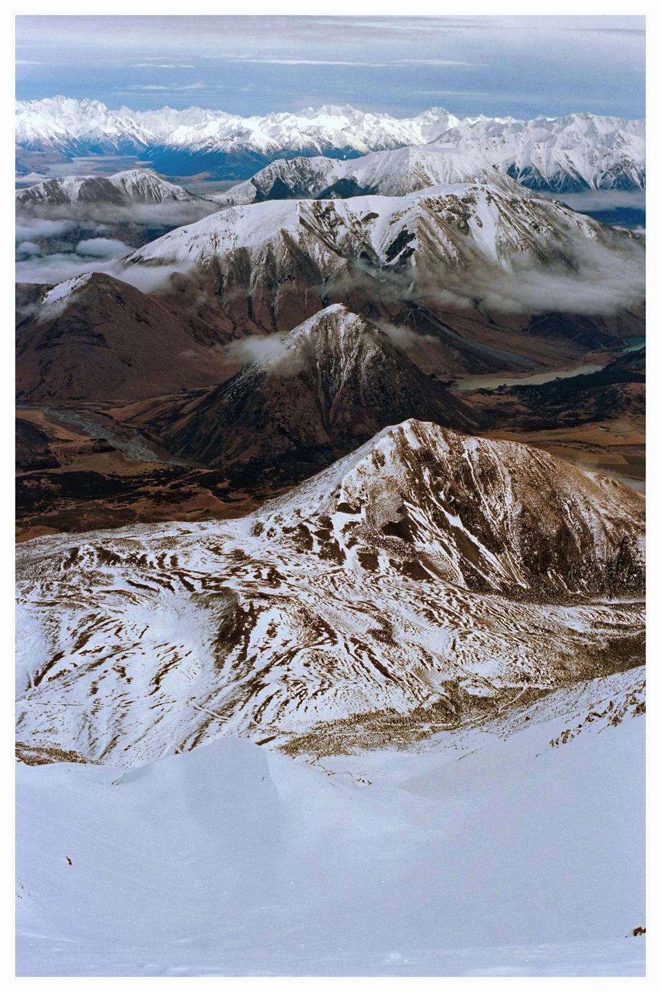 An aerial view shows a vast mountain range with snow-capped peaks and valleys partially obscured by clouds.