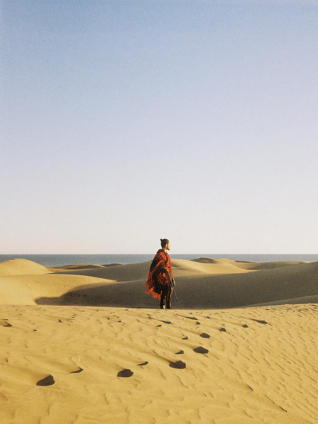 A person wearing a red poncho stands on a sand dune with the ocean in the background.