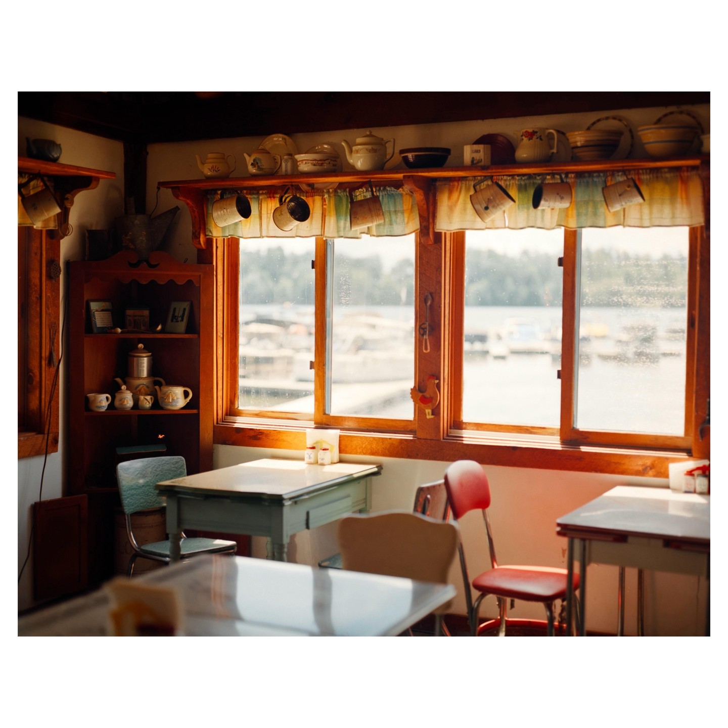 Sunlight streams through a window in a diner, illuminating tables and chairs, with a view of a body of water and boats outside.