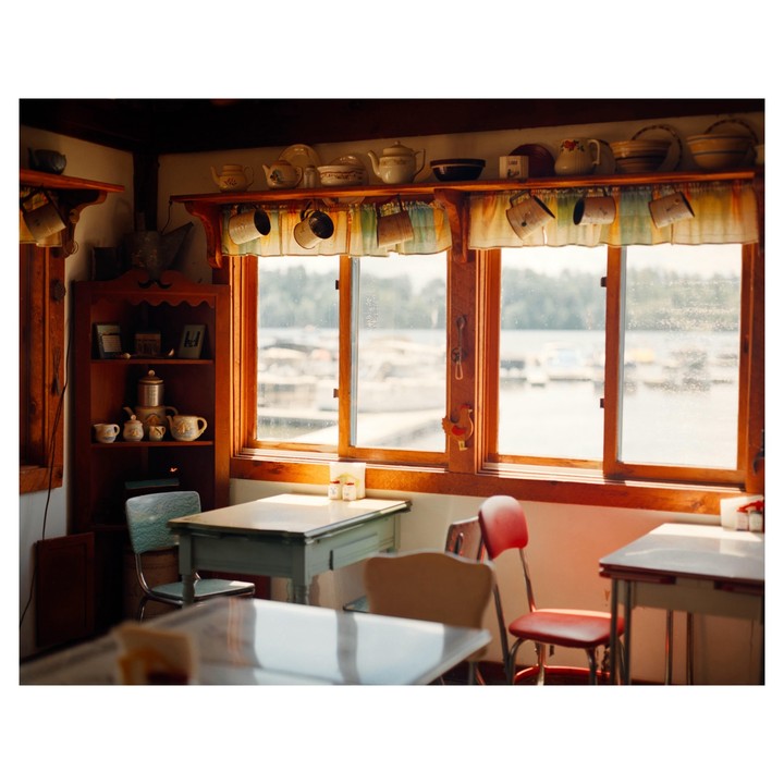 Sunlight streams through a window in a diner, illuminating tables and chairs, with a view of a body of water and boats outside.