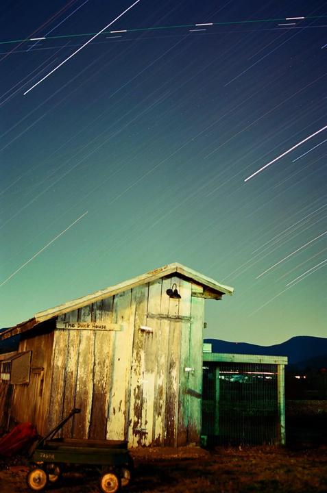 A wooden shed with peeling paint stands under a night sky filled with star trails.