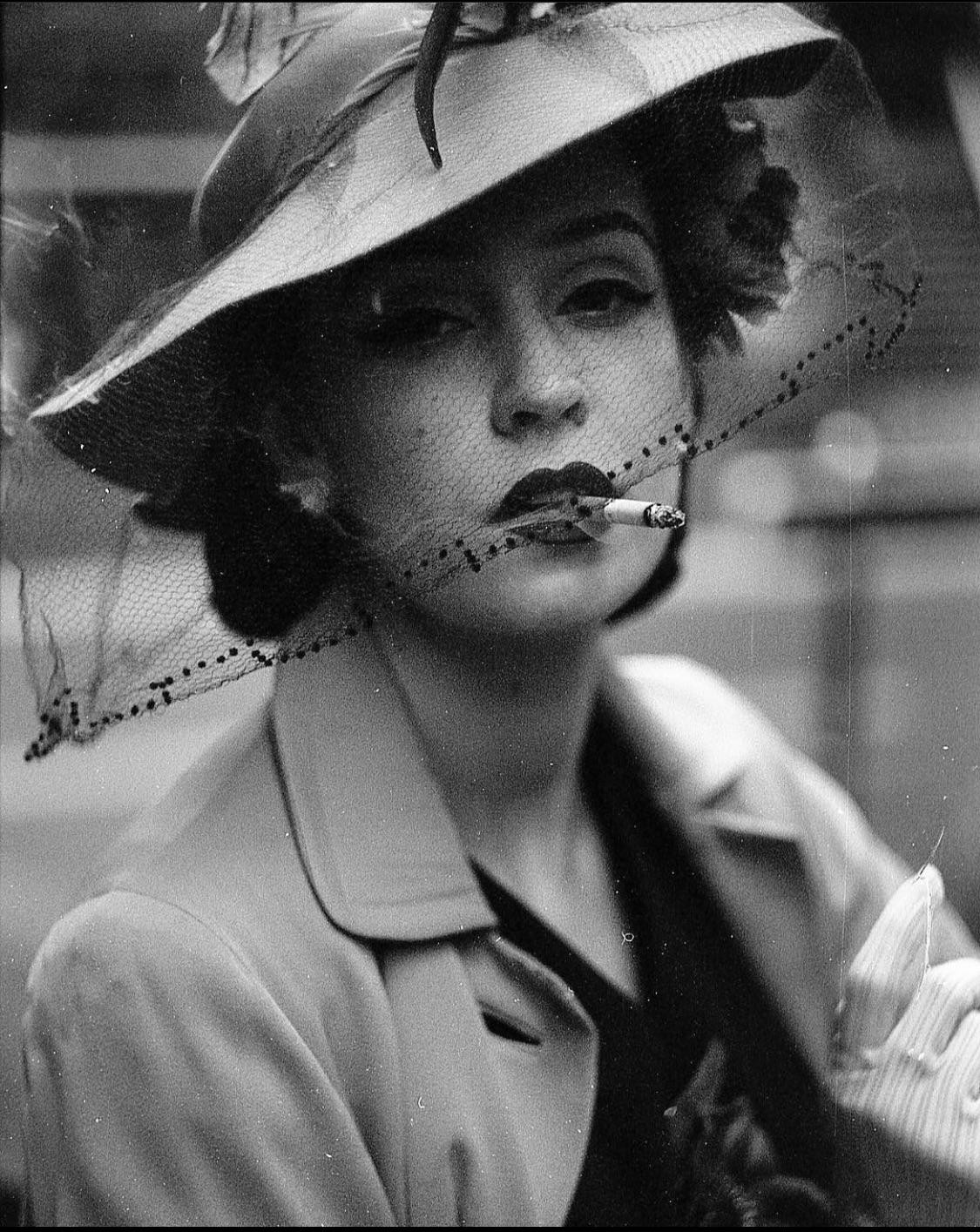 A woman wearing a hat with a veil and a coat holds a cigarette in her mouth.