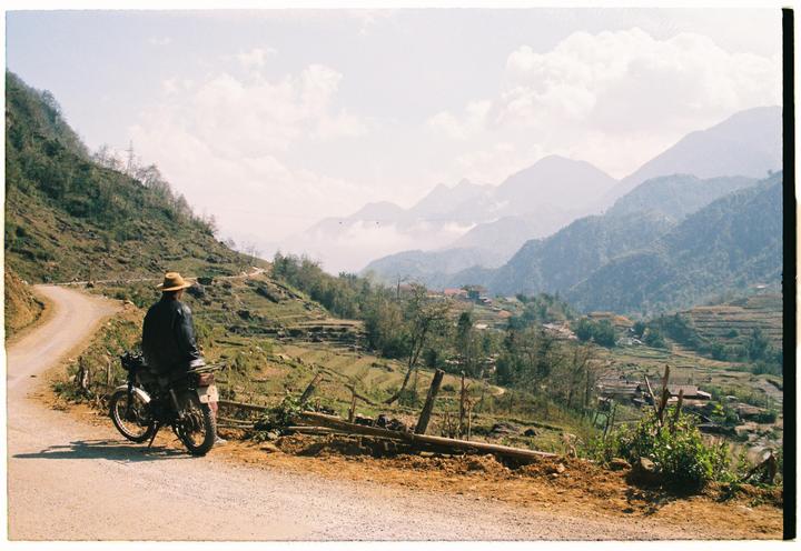A man on a motorcycle looks out over a valley with terraced fields and mountains in the distance.