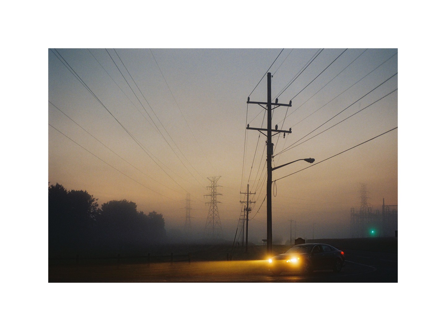 A car with bright headlights drives on a foggy road past power lines and transmission towers at dawn.