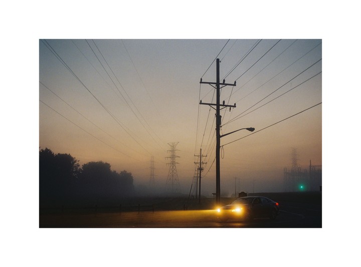 A car with bright headlights drives on a foggy road past power lines and transmission towers at dawn.
