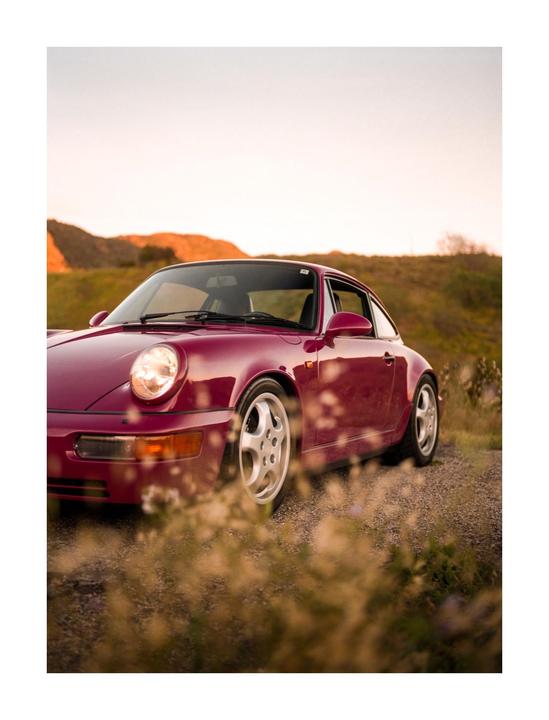 A pink car is parked on a gravel road with hills in the background.