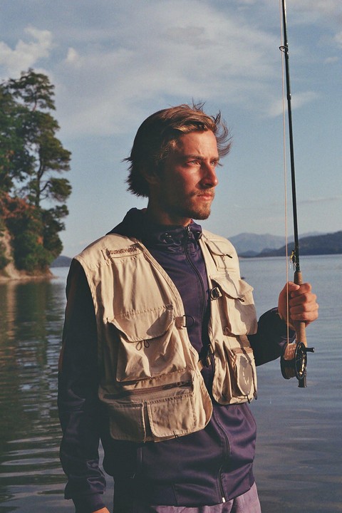A man wearing a fishing vest and holding a fishing rod stands in water with a tree-lined shore and mountains in the background.