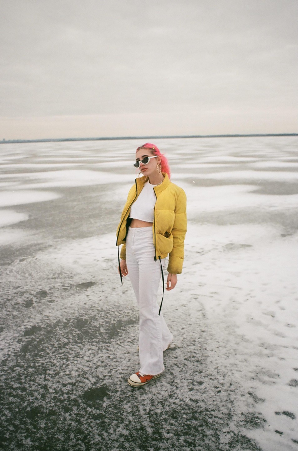 A woman with pink hair, sunglasses, a yellow puffer jacket, and white pants stands on a frozen lake.