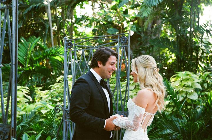A man in a tuxedo and a woman in a white lace wedding dress hold hands and smile at each other in a lush garden.