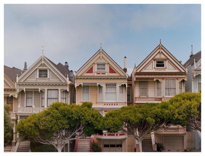 A row of Victorian houses with ornate details and trees in front of them stands under a clear sky.