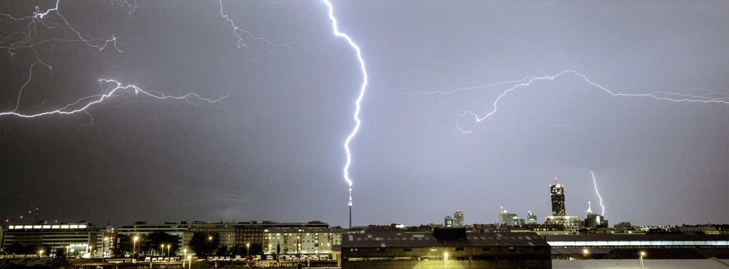 Lightning strikes a tall tower in a city at night, with other buildings and lights visible below a dark, stormy sky.