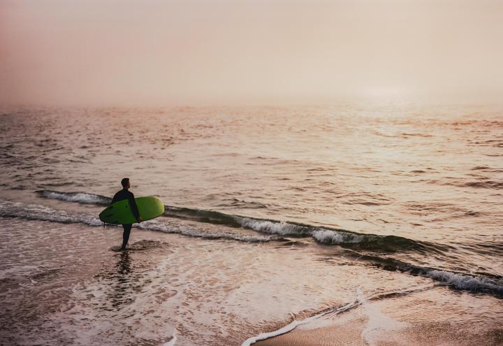 A surfer in a wetsuit stands in the shallow water of a beach, holding a green surfboard under their arm.