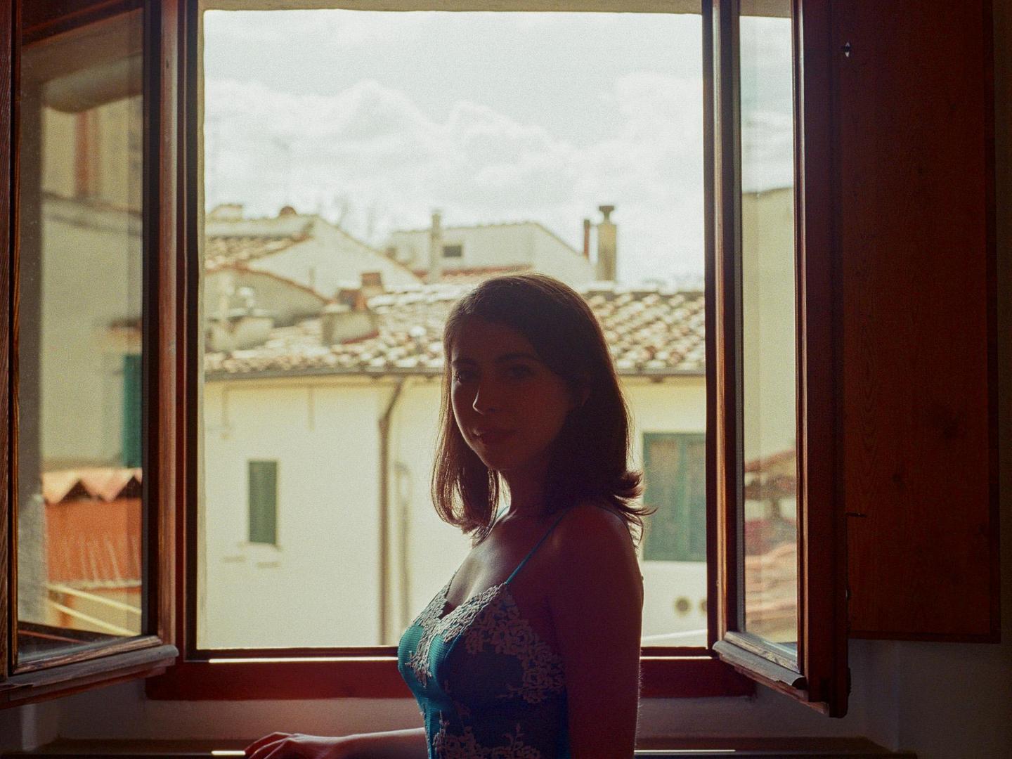 A woman stands in front of an open window looking out at a cityscape with tiled roofs.