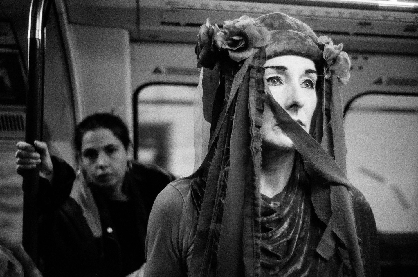 A person with elaborate makeup and a floral headpiece looks up while riding a subway.