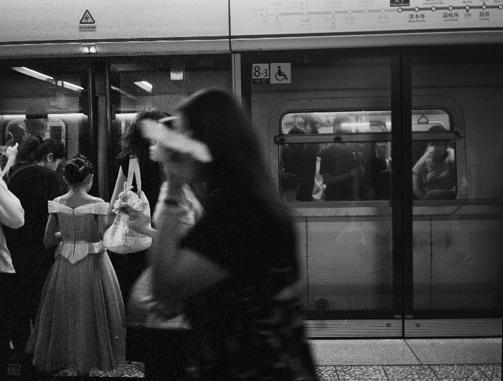 A girl in a dress stands on a subway platform next to a train with people visible through its windows.
