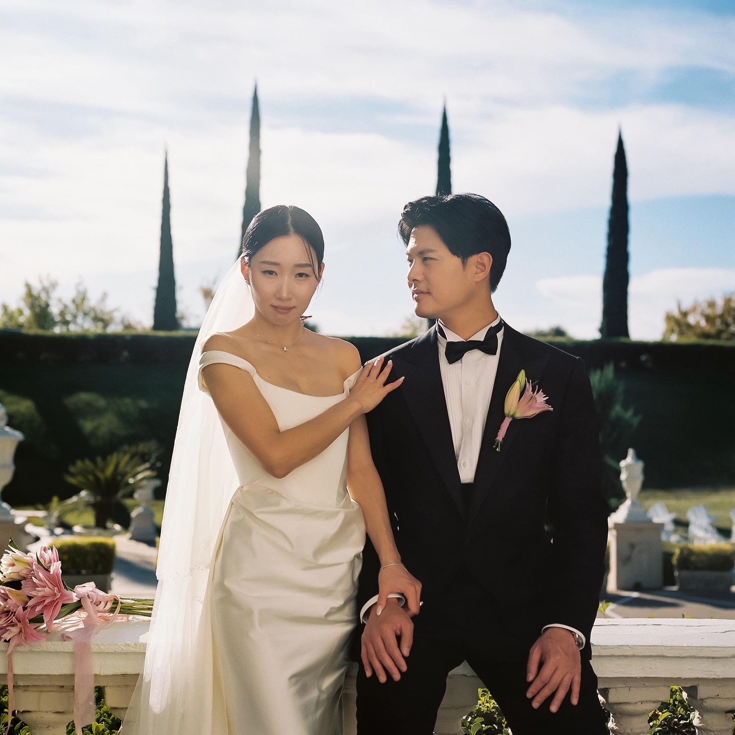 A couple poses for a wedding portrait on a sunny day with tall, narrow trees in the background.