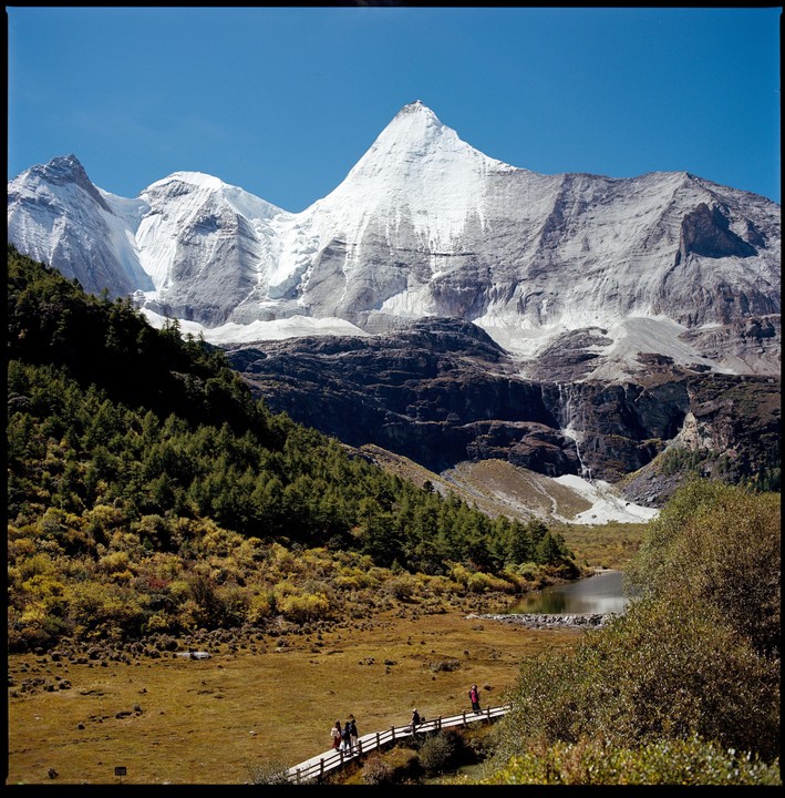 A group of people walk on a wooden boardwalk through a valley with a lake and snow-capped mountains in the background.