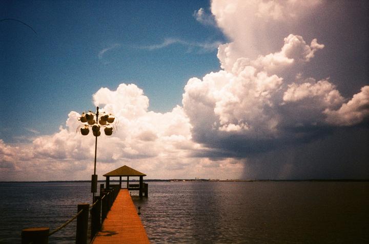 A long wooden pier with a gazebo at its end extends into a body of water under a sky with both white clouds and dark storm clouds.