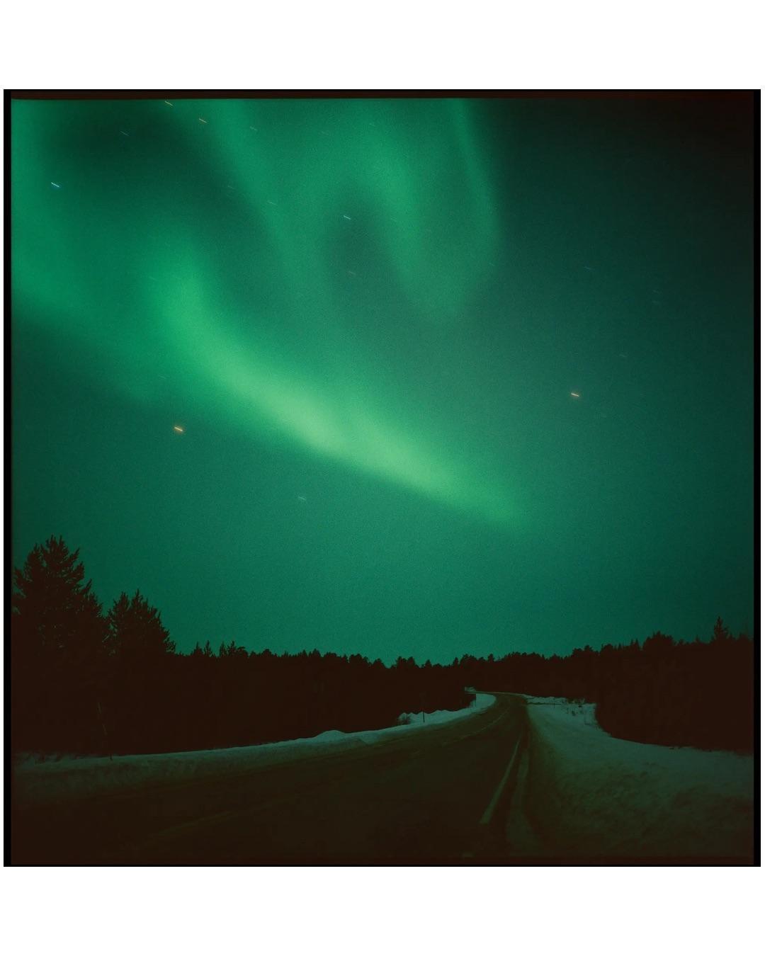 The aurora borealis illuminates a snowy road winding through a forest at night.