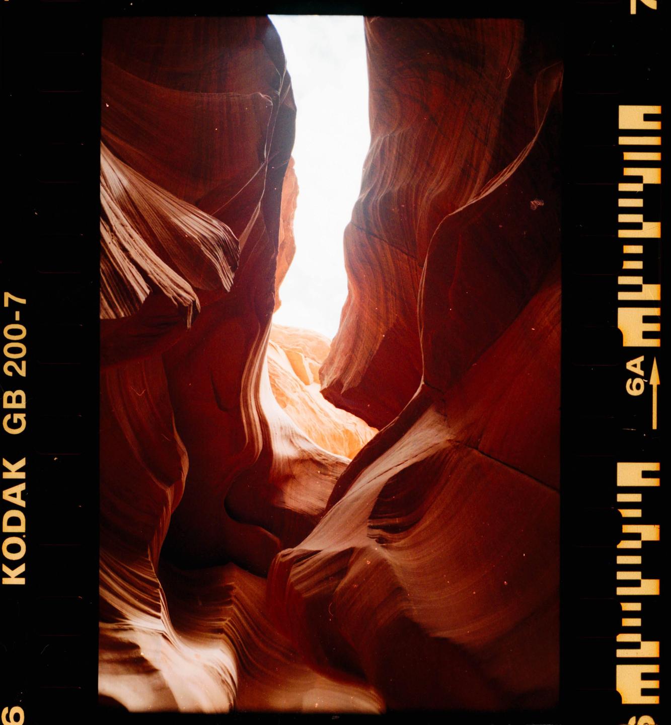 A narrow slot canyon with red and orange rock formations is illuminated by bright light from above.