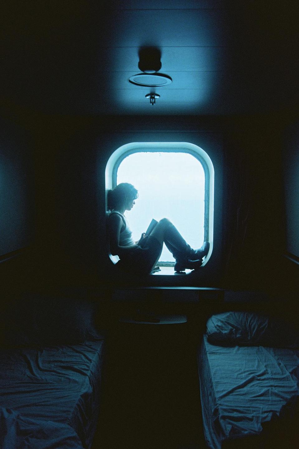 A woman sits in a window reading a book in a dark room with two beds.