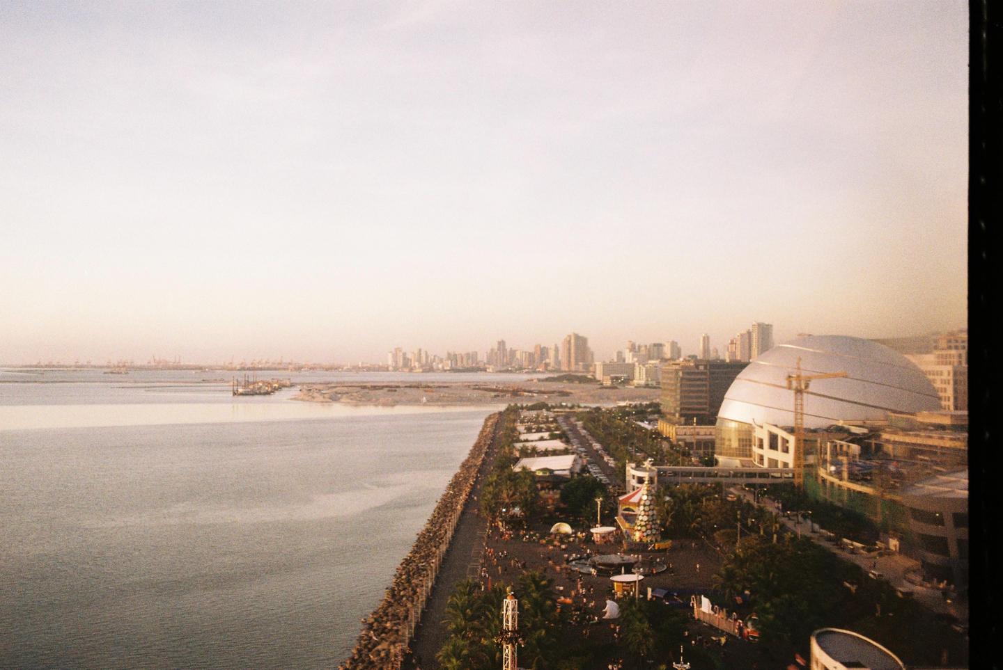 A wide shot shows a city skyline and a large body of water with a rocky shore and a fairground in the foreground.