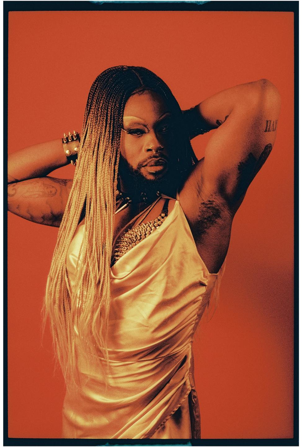 A person with long braided hair and a beard poses against a red background, wearing a gold dress and jewelry.