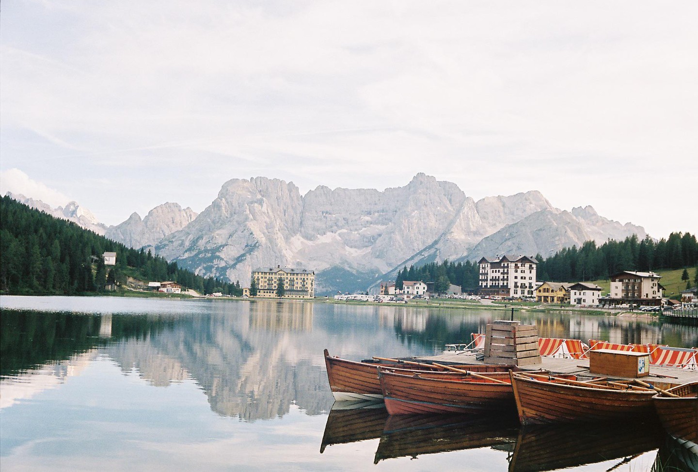 Several wooden boats are docked on a calm lake with mountains and buildings in the background.