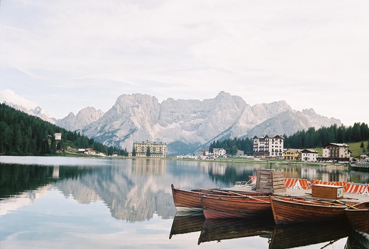 Several wooden boats are docked on a calm lake with mountains and buildings in the background.