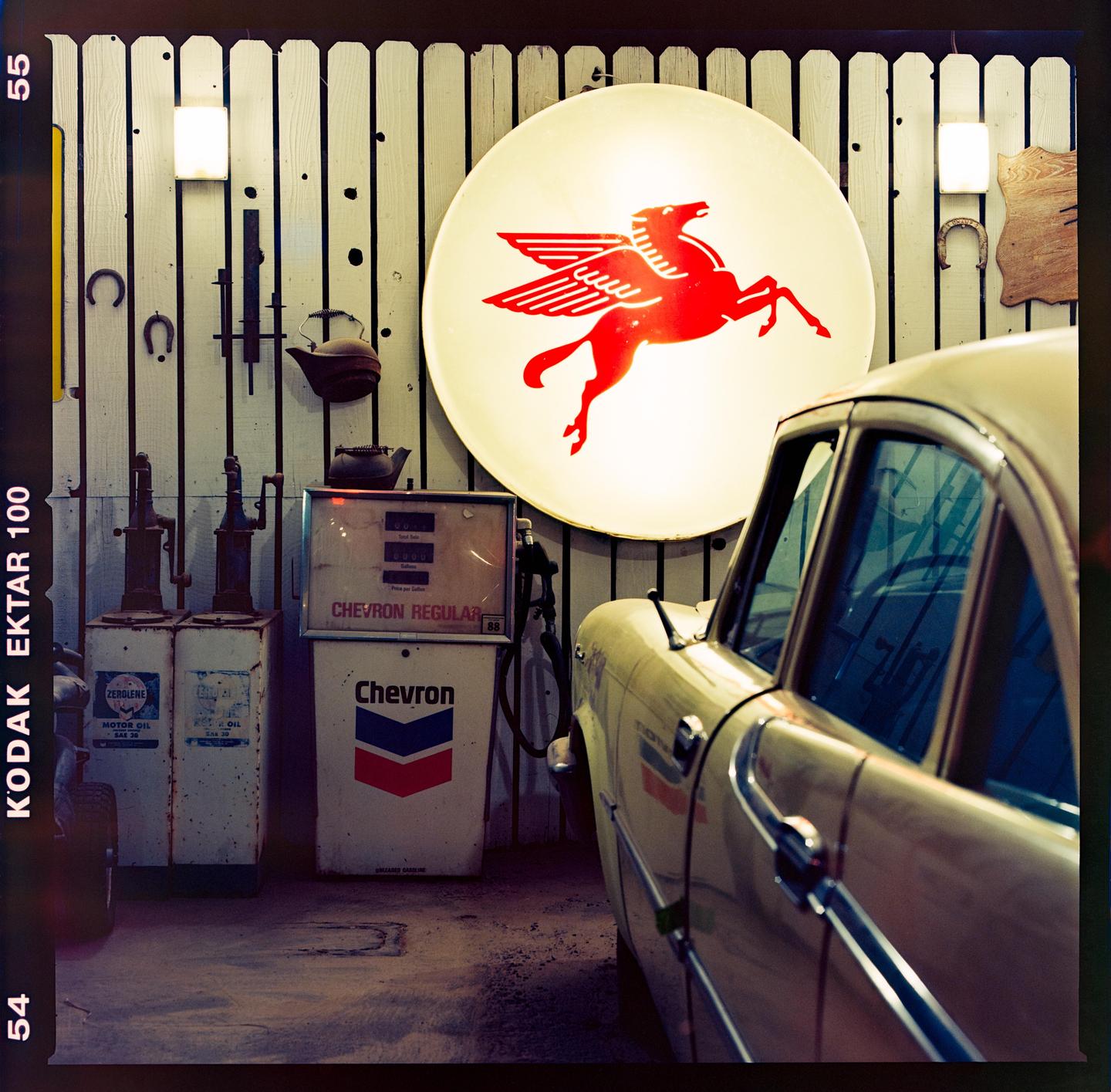 A vintage car is parked next to old gas pumps and a large illuminated sign with a red Pegasus logo.