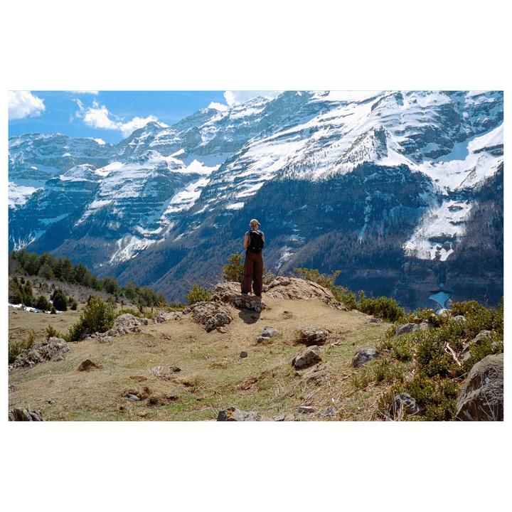 A woman with a backpack stands on a rocky outcrop overlooking a valley with snow-capped mountains.