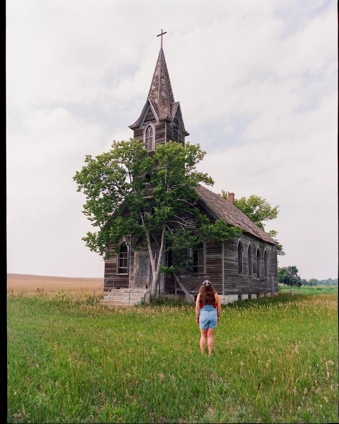 A woman stands in a field of tall grass, looking at an old wooden church with a tall steeple.