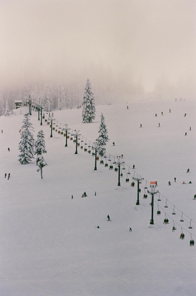 A ski slope with a chairlift and many skiers is covered in snow, with snow-covered trees and a hazy sky in the background.