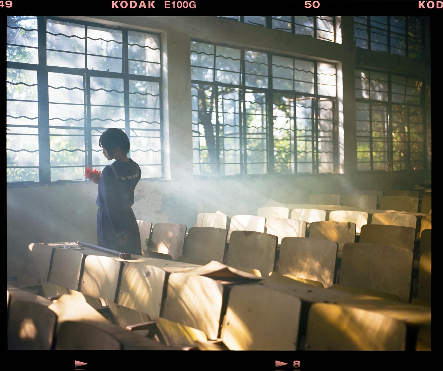 A girl in a school uniform stands in a dusty classroom holding a red flower, with light rays shining through the windows.