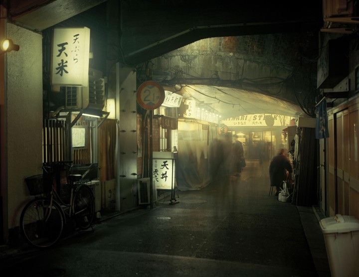 A bicycle is parked next to a building with illuminated signs in a narrow underpass where people are walking.
