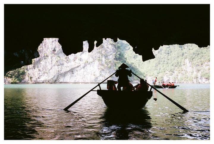 A person in a conical hat rows a boat with passengers out of a dark cave toward a body of water with limestone cliffs and other boats.