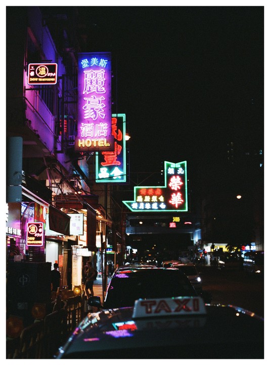 A street at night is filled with neon signs and a taxi is parked in the foreground.