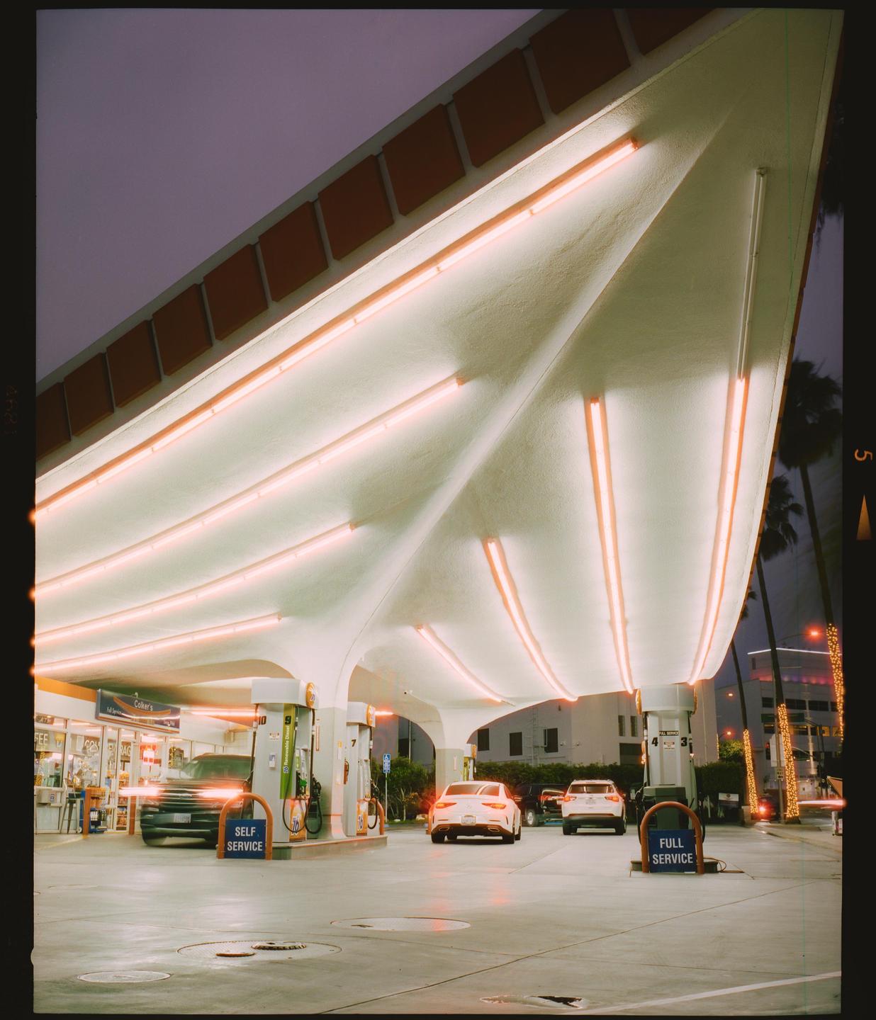 A gas station with a distinctive roof design is illuminated by neon lights at dusk, with several cars parked at the pumps.