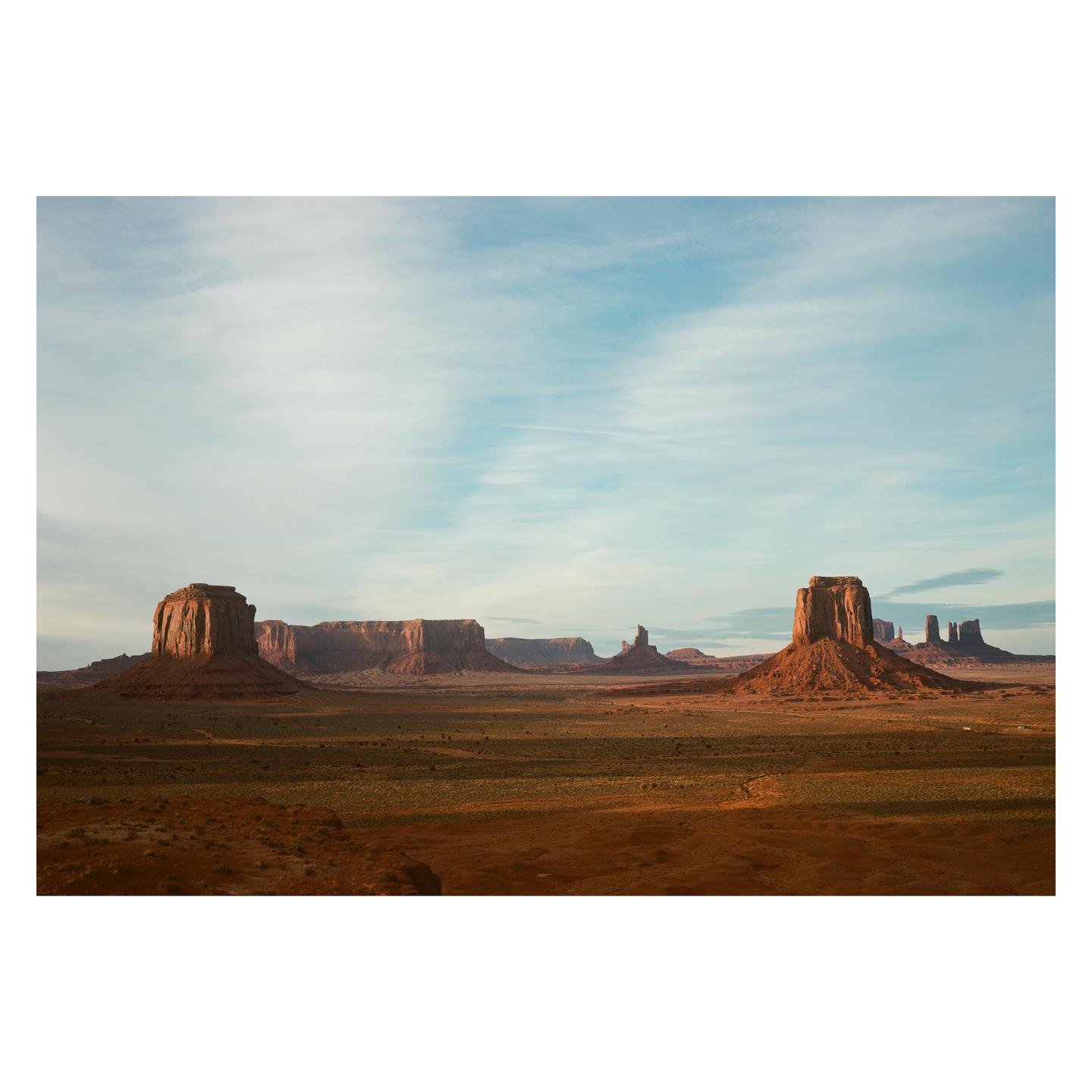 A wide landscape shows several large rock formations rising from a flat, arid plain under a partly cloudy sky.