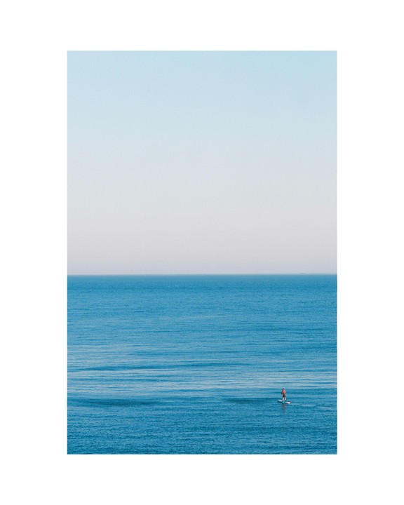 A person on a paddleboard floats on a calm blue ocean under a clear sky.