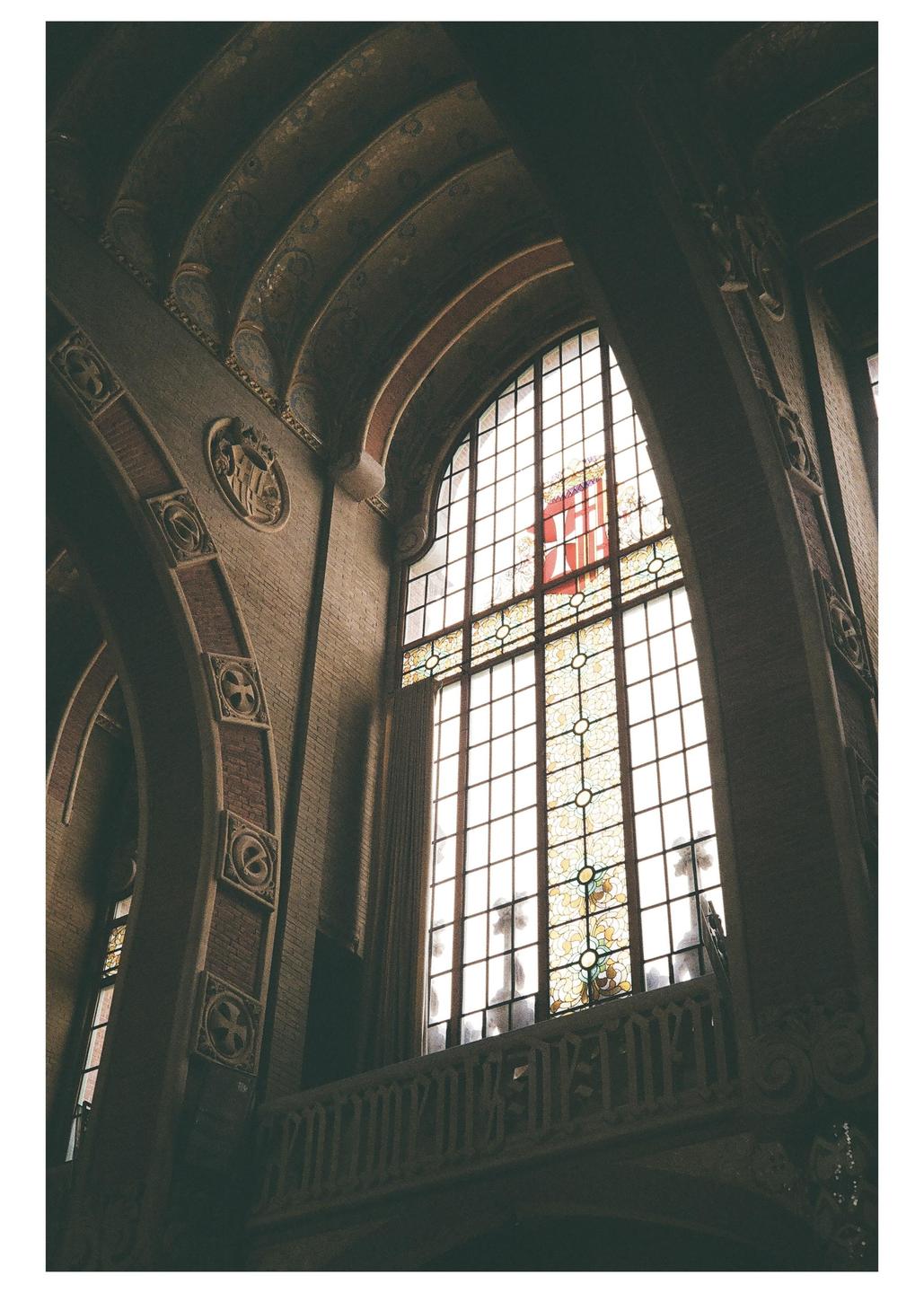A large stained glass window with a red emblem is visible through an arched opening in a brick building.