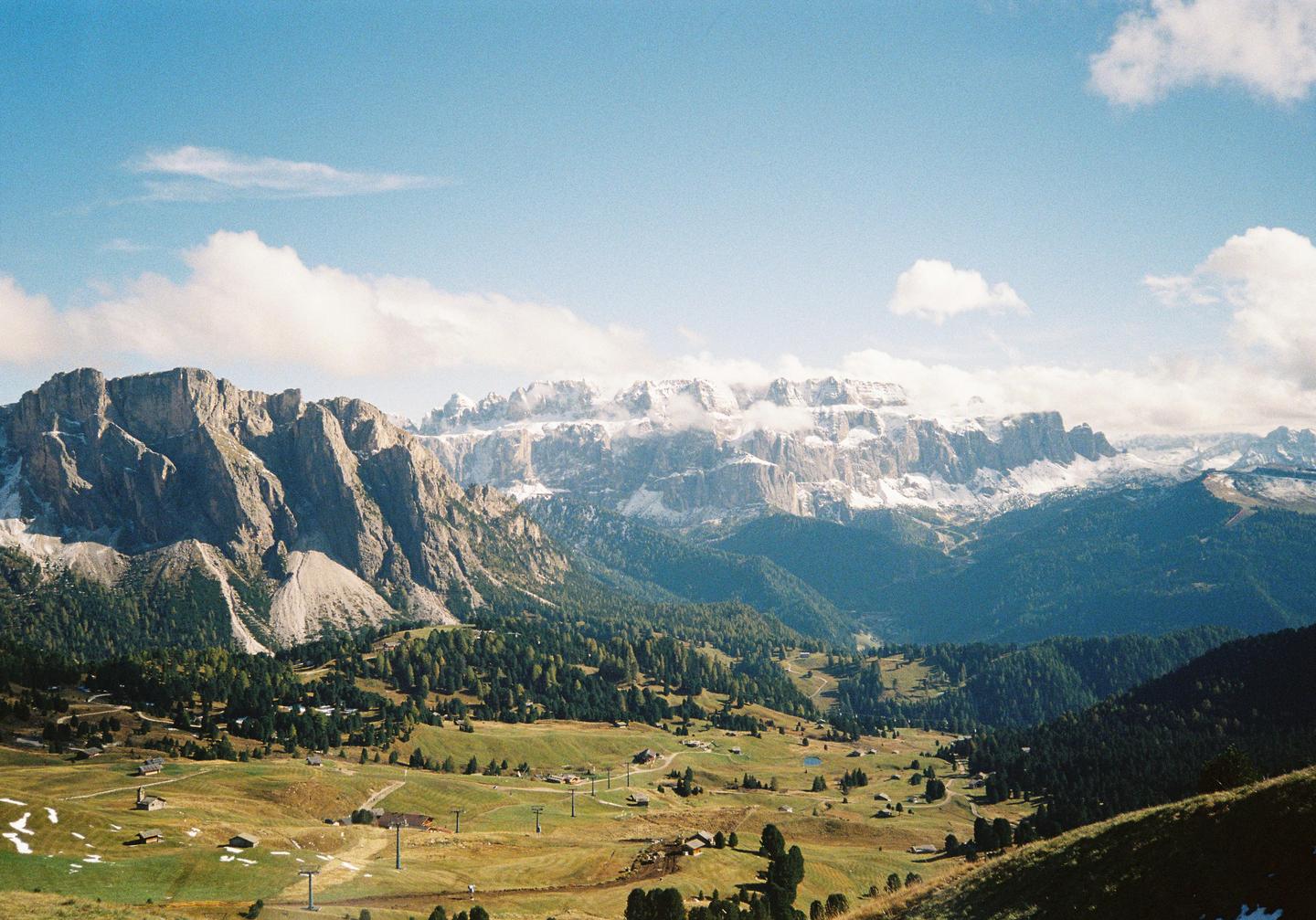 A vast mountain landscape features a valley with scattered buildings and trees, leading up to a range of rocky peaks, some of which are snow-capped.
