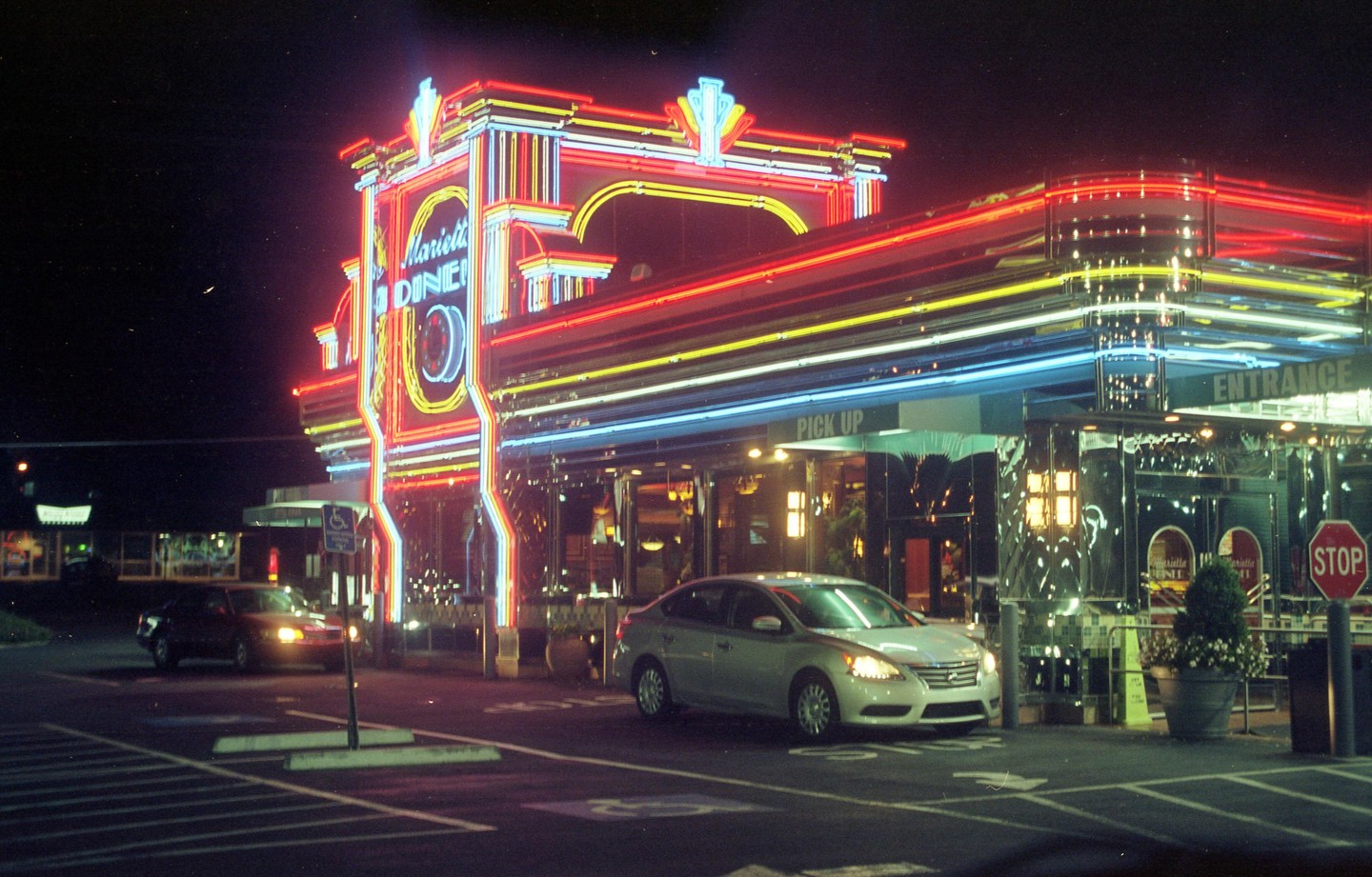 A diner with bright neon lights and two cars in the parking lot is visible at night.