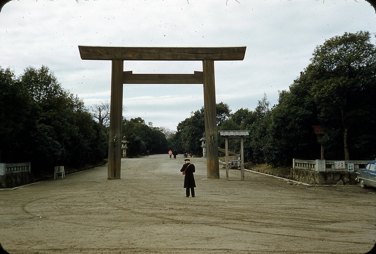 A man in a uniform stands in the middle of a wide dirt path leading to a large wooden torii gate, with trees and stone lanterns on either side.