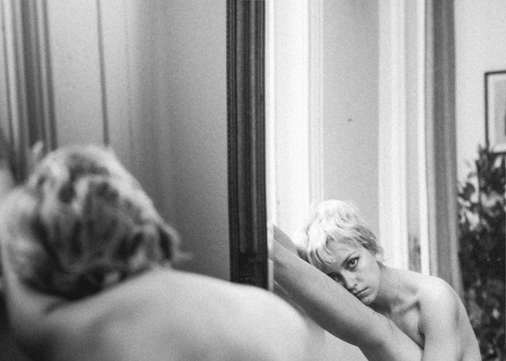 A woman with short, light hair looks into a mirror, with her reflection visible in the foreground.