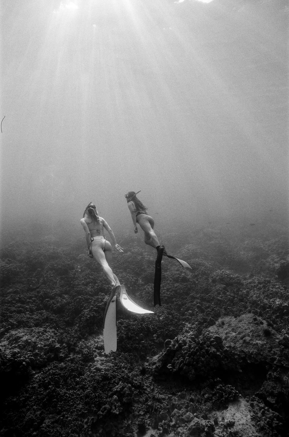 Two women wearing fins and swimsuits swim underwater above a rocky seabed with sun rays shining through the water.