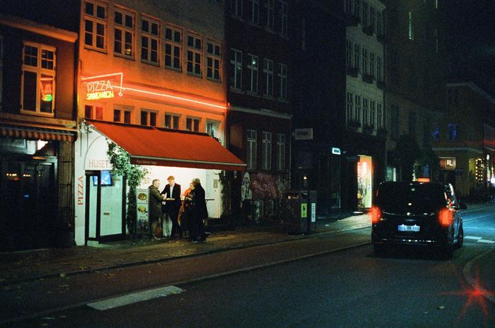 A group of people stand outside a pizza shop with a red awning and neon sign at night.