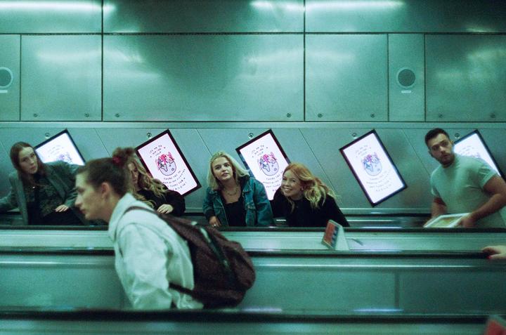 Several people are riding an escalator in a subway station with illuminated advertisements on the wall.